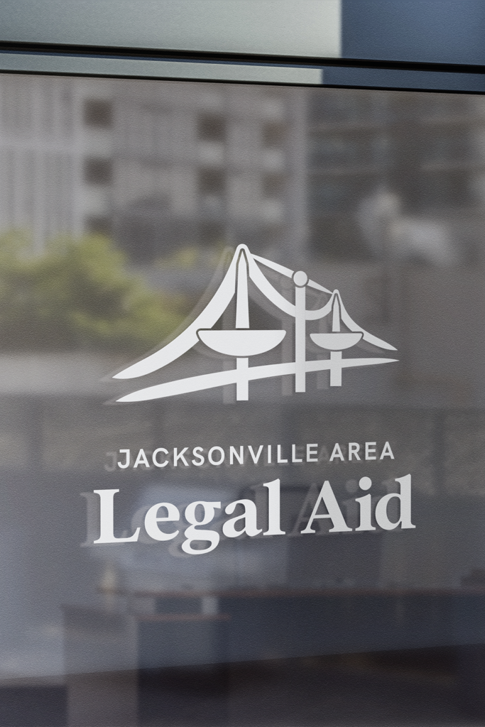 Close-up of a glass door or window with the Jacksonville Area Legal Aid logo and name in white. The logo depicts a stylized bridge with scales of justice. A blurred urban scene is visible in the reflection.