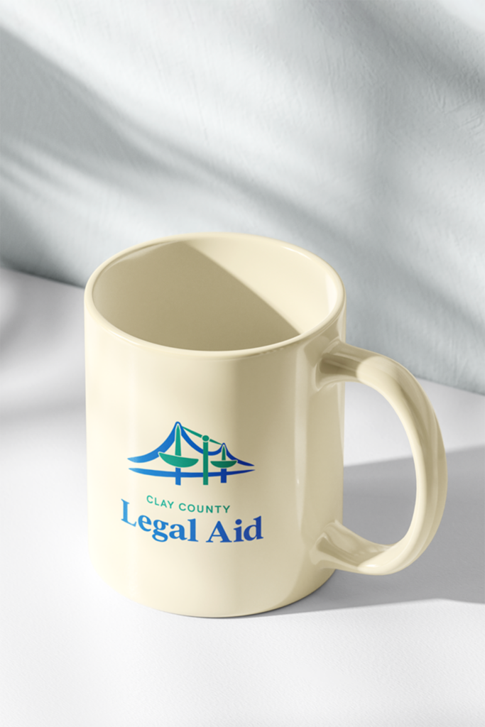 A light yellow coffee mug with the Clay County Legal Aid logo and text printed in blue and green, sitting on a white table.